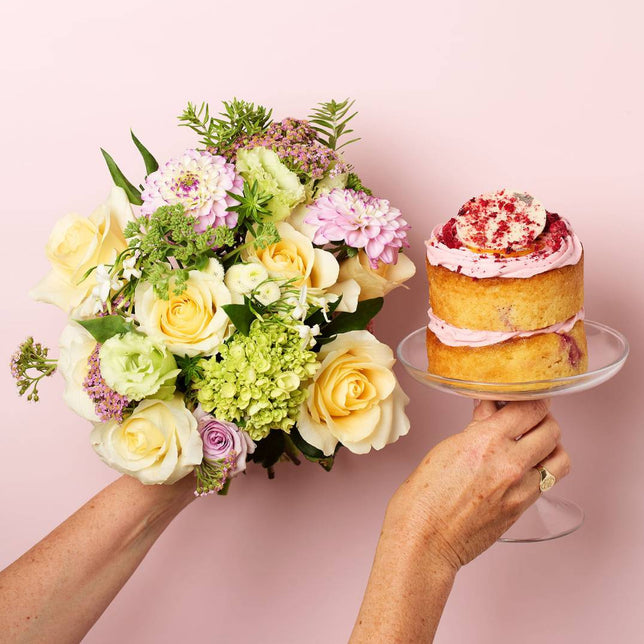 Two hands holding a bouquet of flowers and a small cake on a pink background