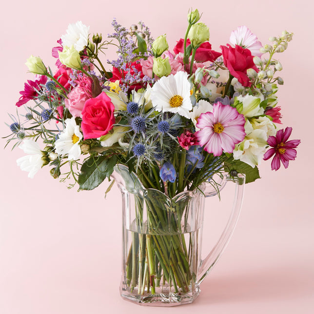 A colorful fresh flower bouquet arranged in a clear glass vase, with a variety of flowers including roses and daisies, on a pink background.