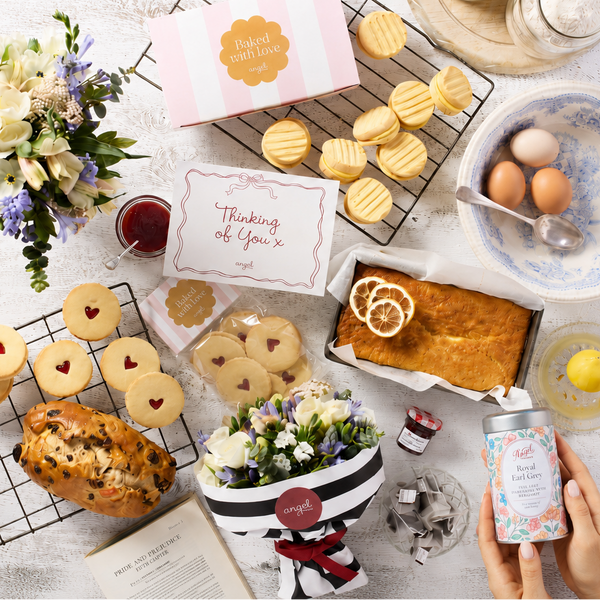A styled flat-lay of baked goods and tea accessories on a white wooden surface, with a hand holding a can of tea in the lower right corner. Visible items include assorted cookies, a lemon cake, flowers, a bowl with eggs, and a pink and white box.