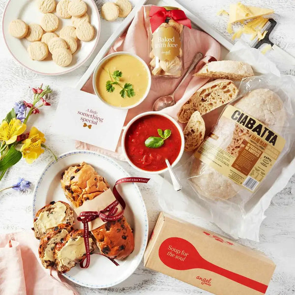 Soup and bread hamper with ciabatta loaf, hot cross buns, cookies, two soups, and a gift card arranged with flowers.