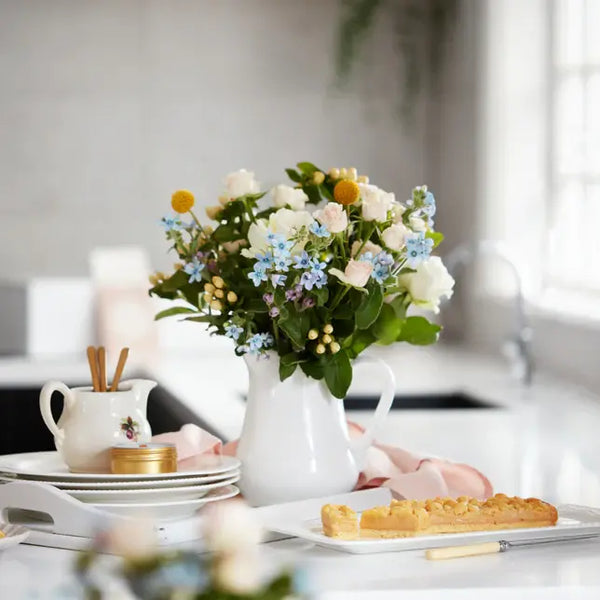 White jug with pastel floral bouquet on a kitchen counter beside plates and dessert