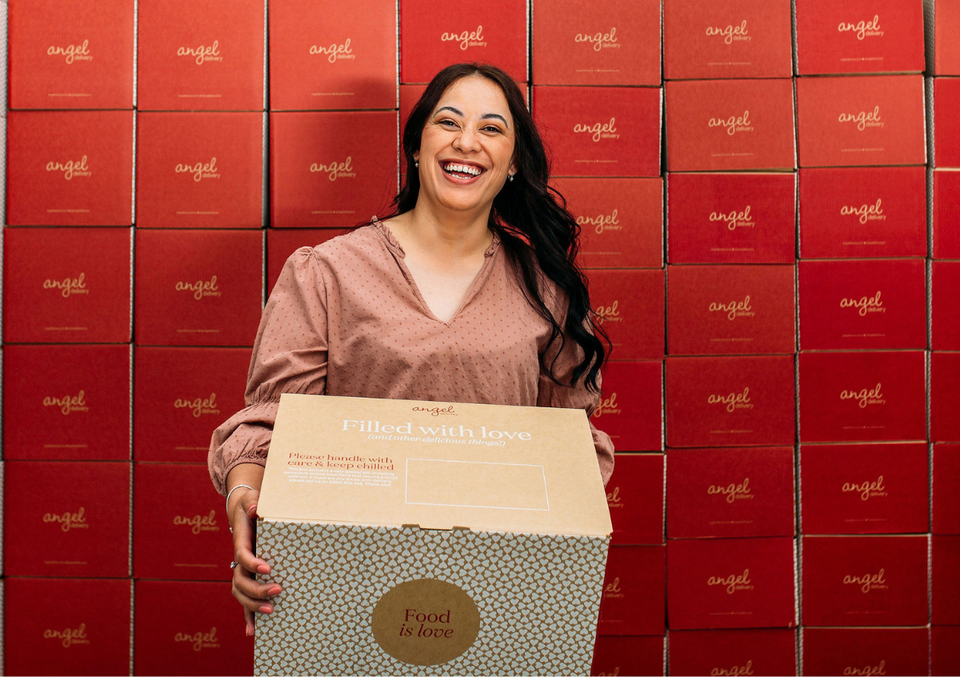 Smiling woman holding Angel Delivery box in front of stacked red branded boxes