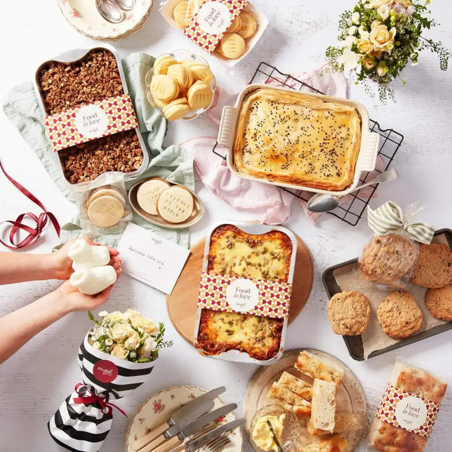 Food hamper with lasagna, pie, cookies, granola, shortbread, bread, and flowers arranged on a table with a gift card.