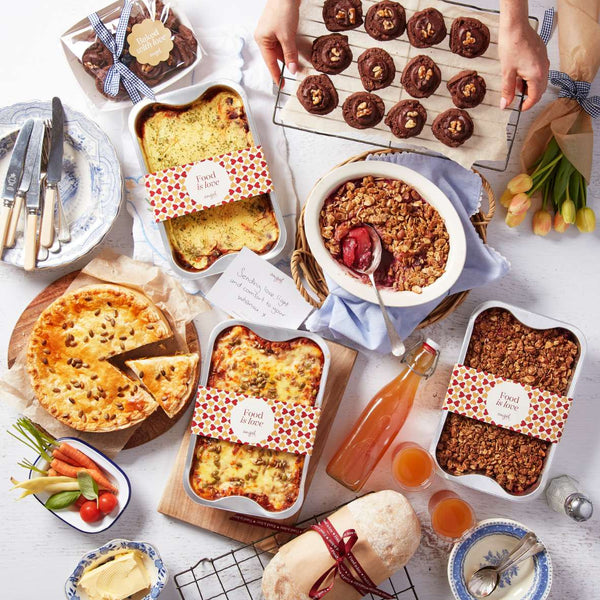 Assorted meal hamper with lasagna, pie, granola, bread, juice, cookies, and vegetables arranged on a table with a gift card.