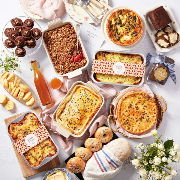 Assorted food hamper with quiches, lasagna, bread rolls, muffins, crumble, juice, and baked treats arranged on a table.