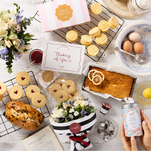 A styled flat-lay of baked goods and tea accessories on a white wooden surface, with a hand holding a can of tea in the lower right corner. Visible items include assorted cookies, a lemon cake, flowers, a bowl with eggs, and a pink and white box.